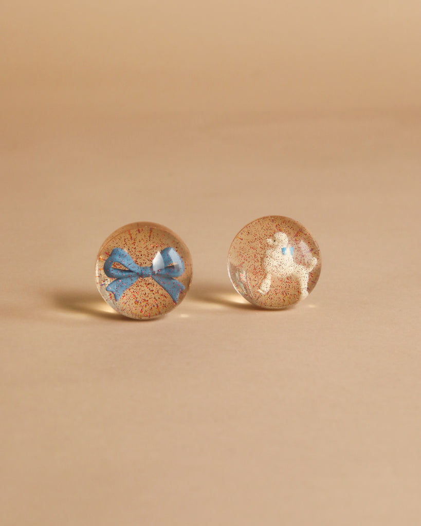 Two clear, round Konges Sloejd Glitter Bouncing Balls—one with a tiny blue bow inside, the other featuring red glitter specks—rest on a beige surface. Set name: Poodle & Bow.