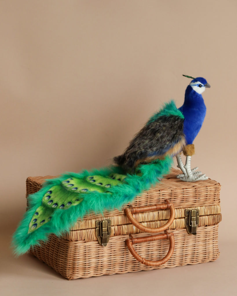 A vibrant Peacock Stuffed Animal with striking blue and green feathers, perched on a woven wicker suitcase against a soft beige background.