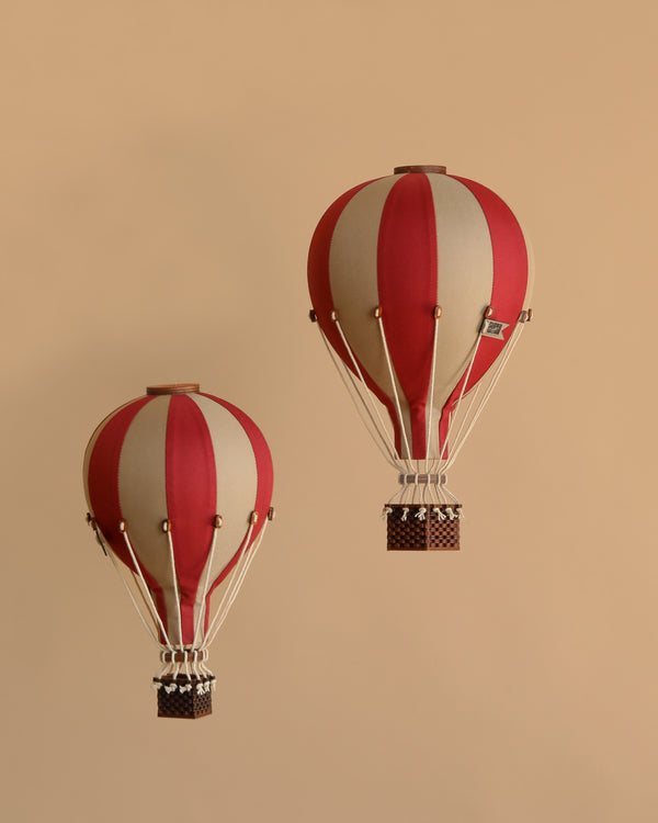 Two burgundy and white striped handmade Hot Air Balloon - Burgundy decorations with wicker baskets float against a beige background. Vintage-inspired, they add whimsical charm to any children's room or space.