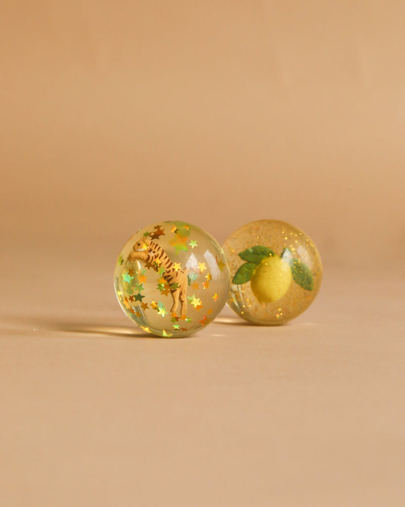 Two transparent bouncing balls from the Konges Sloejd Glitter Bouncing Ball Set—Lemon & Tiger—rest on a beige surface: one showcases a playful lemon with glitter, while the other features a tiger design with sparkling confetti inside.