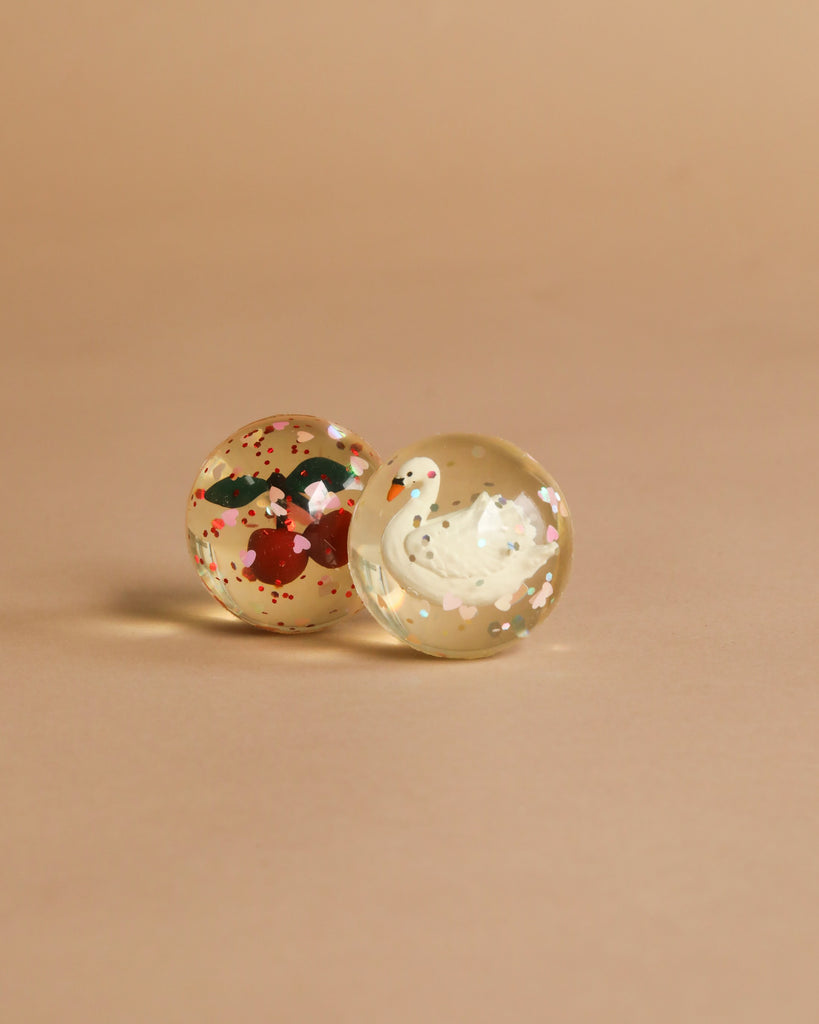 The Konges Sloejd Glitter Bouncing Ball Set - Cherry & Swan includes two transparent balls, one with a swan and one with red cherries and green leaves, both filled with glitter and colorful flecks for playful fun.