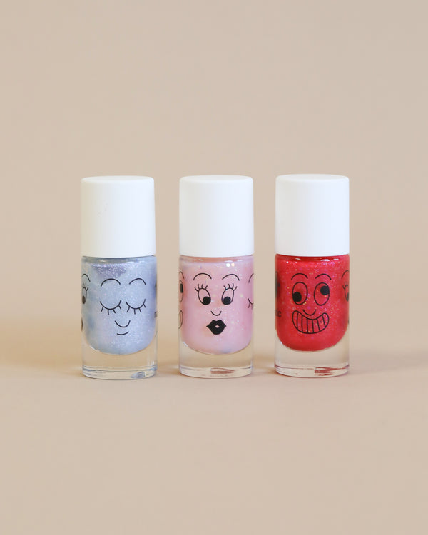 Three colorful water-based Nailmatic - 3 Nail Polish Set - Mermaid bottles with cartoon faces drawn on them; one blue, one pink, and one red, lined up against a soft beige background.