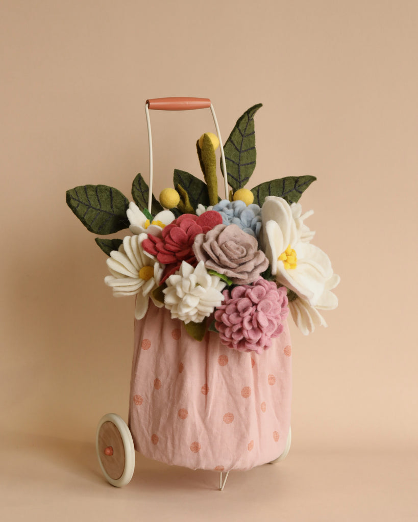 A small peach-colored cart with white polka dots holds a bouquet of handmade fabric flowers. The bouquet includes white daisies, pink and purple roses, red flowers, and green leaves. The Felt Flower Trolley Basket has white wheels and a peach-colored handle against a beige background, showcasing eco-friendly craftsmanship.