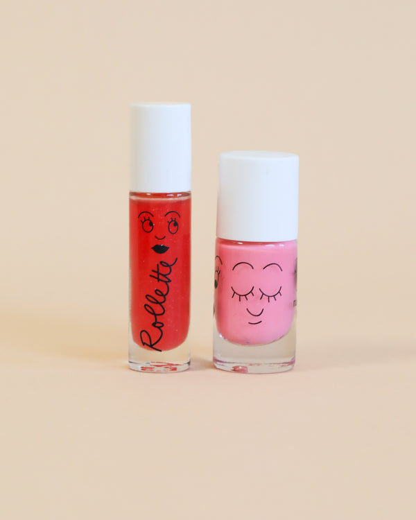 Two bottles of Nailmatic water-based nail polish with cute faces drawn on them, one pink and one red, against a soft peach background.