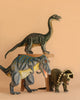 Three Giganotosaurus Dinosaur Stuffed Animals are arranged against a beige background. One sits atop a small wooden stool, while the other two are placed in front of it on the ground.