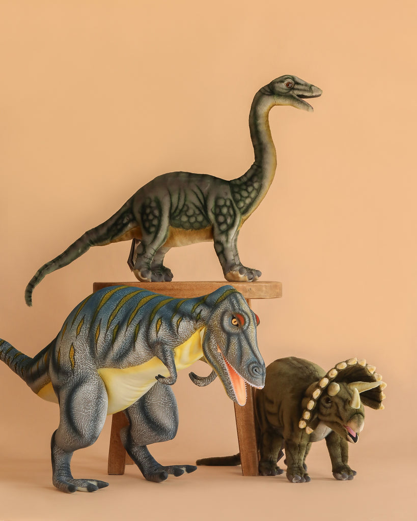 Three Giganotosaurus Dinosaur Stuffed Animals are arranged against a beige background. One sits atop a small wooden stool, while the other two are placed in front of it on the ground.