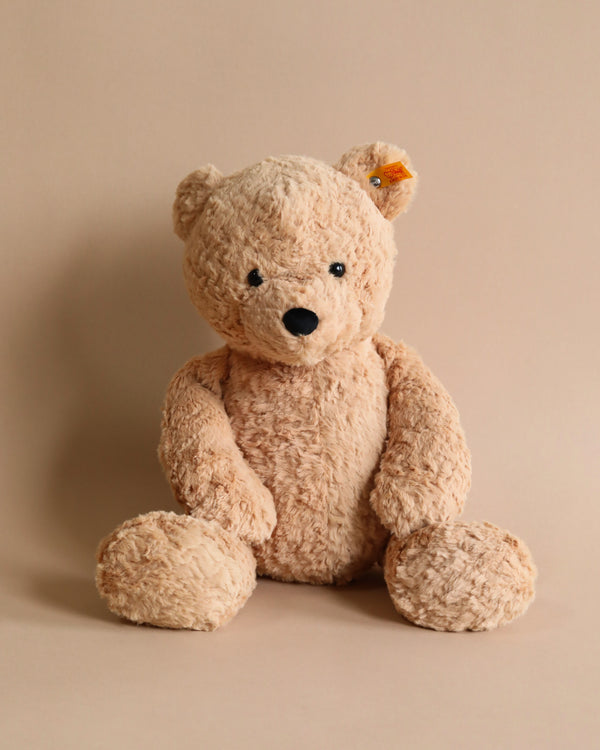 A Steiff Jimmy Teddy Bear, 16 Inches with a light brown, textured fur and a black nose, sits against a plain beige background. It has an alert expression with its head slightly tilted to one side and sports the distinctive