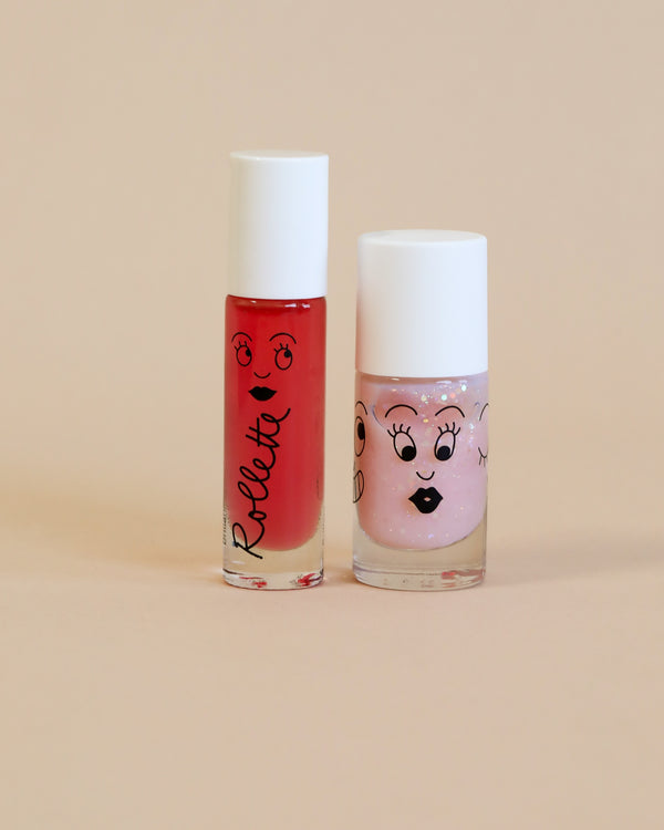 Two Nailmatic - Nail Polish + Lipgloss Set - Fairytails tubes against a beige background; one is red with the word "roller" and smiley faces, and the other is sparkly pink with a cartoon face.