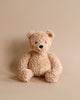 A fuzzy, light brown Steiff Jimmy Teddy Bear sitting upright against a plain, beige background. The bear has black eyes, a shiny nose, and a gently smiling expression.