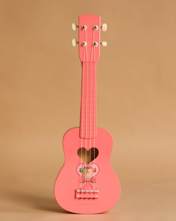 The Konges Sloejd Wooden Ukulele - Bow Kitty features a pink finish, heart-shaped sound hole, and a butterfly design below the strings. This charming instrument stands upright against a plain beige background.
