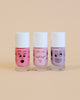 Three bottles of Nailmatic water-based nail polishes with cartoon faces drawn on them, in shades of pink and purple, against a pale yellow background.