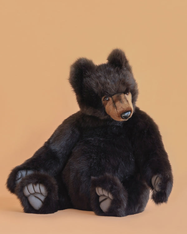 The Brown Tommy Bear Stuffed Animal, 30", features soft dark brown fur, large visible paws, and realistic detailing. It is pictured sitting on a light peach background with its head slightly tilted to the side.