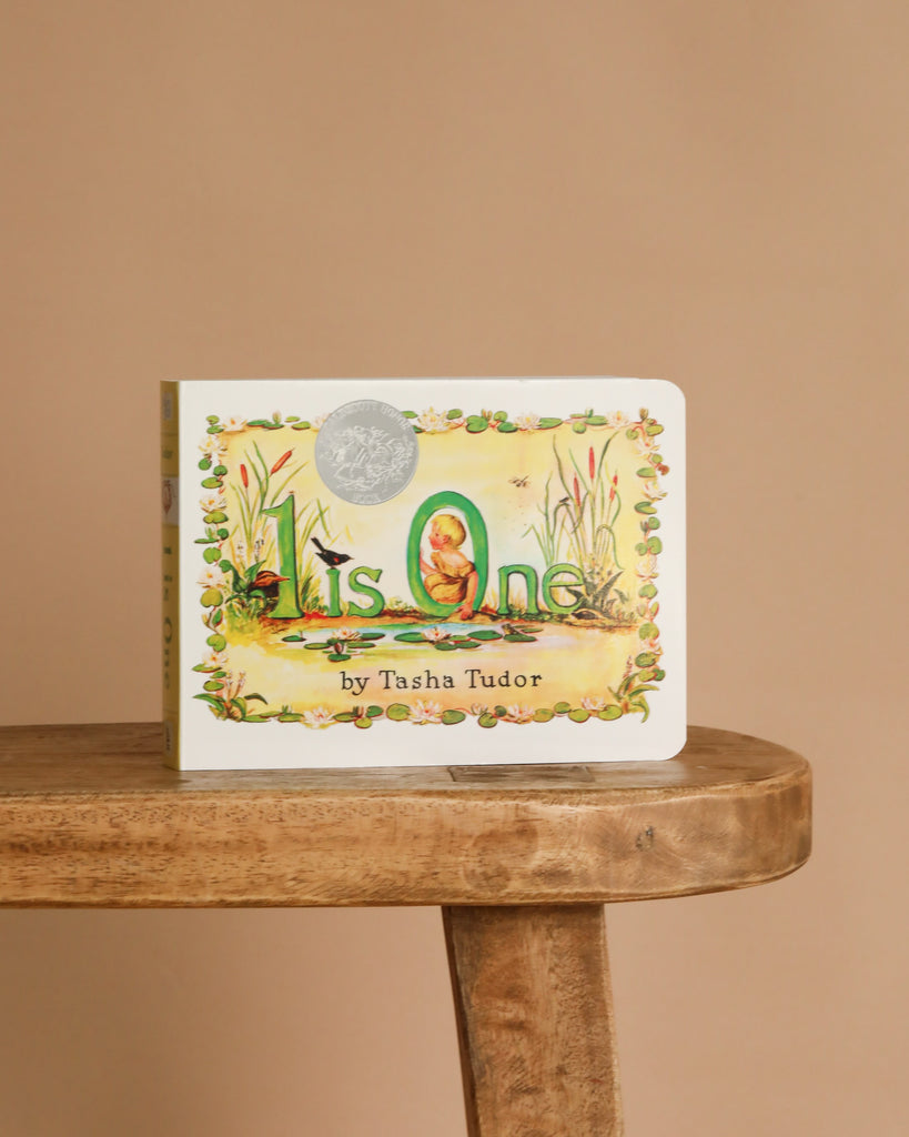 1 Is One by Tasha Tudor" rests on a wooden bench with a plain beige backdrop. Its cover shows a child among flowers and animals, making it an ideal addition to any counting book collection.