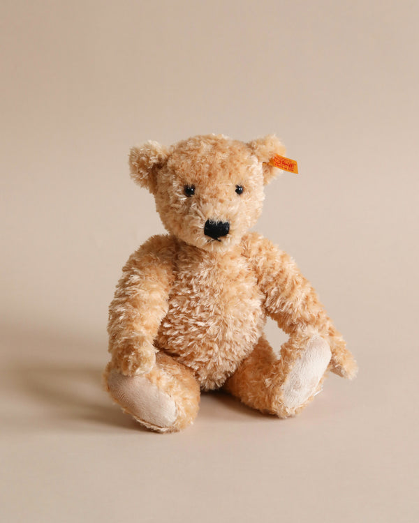 A soft, fluffy Steiff, Elmar Teddy Bear with beige fur sitting against a plain, light brown background. The bear has shiny black eyes and a prominent black nose, with a small, beige button in the ear.