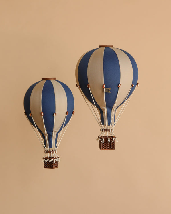 The Hot Air Balloon - Navy Blue features blue and beige vertical stripes with a woven basket, displayed against a plain beige background. This handmade piece is perfect as a charming children's room decoration or unique accent.
