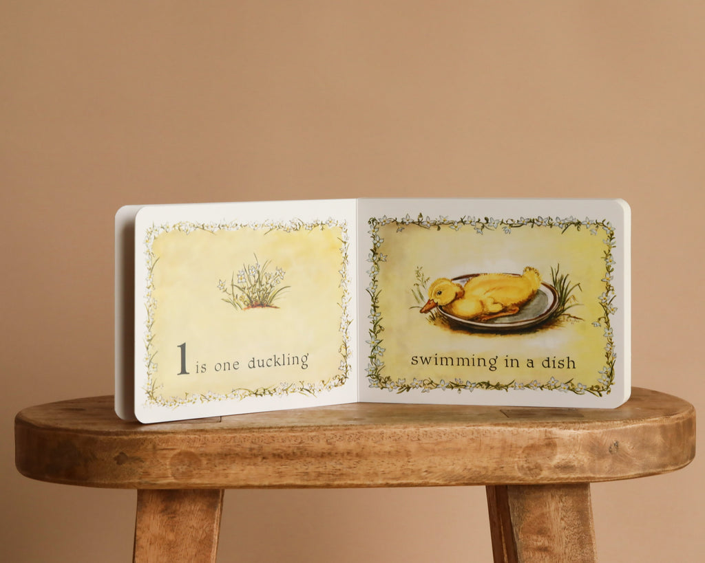 An open copy of "1 Is One by Tasha Tudor" sits on a wooden stool, displaying "1 is one duckling" with a duckling swimming in a dish—an ideal first counting book for little readers.