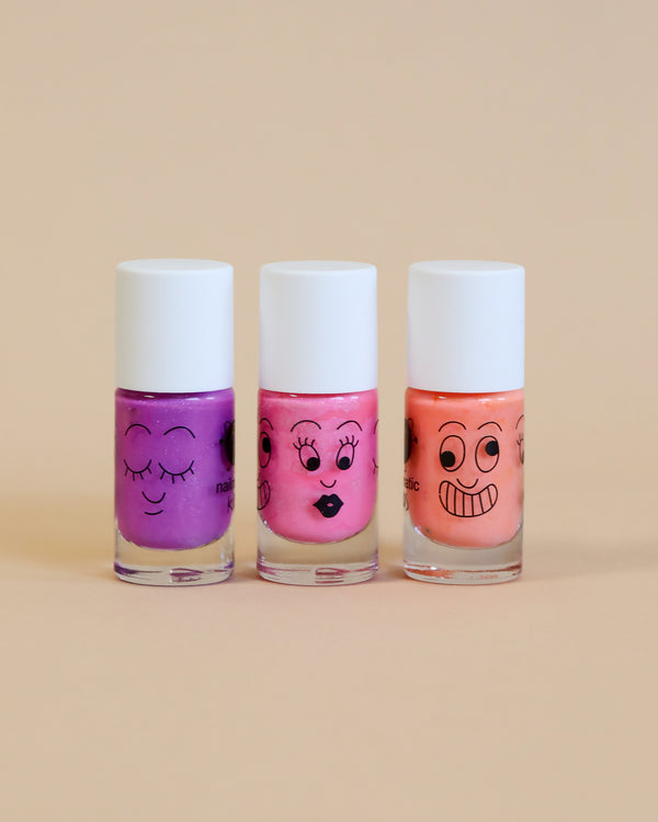 Three bottles of Nailmatic - 3 Nail Polish Set - Paris with cartoon faces, arranged in a row against a soft beige background. The colors are purple, pink, and peach from left to right.
