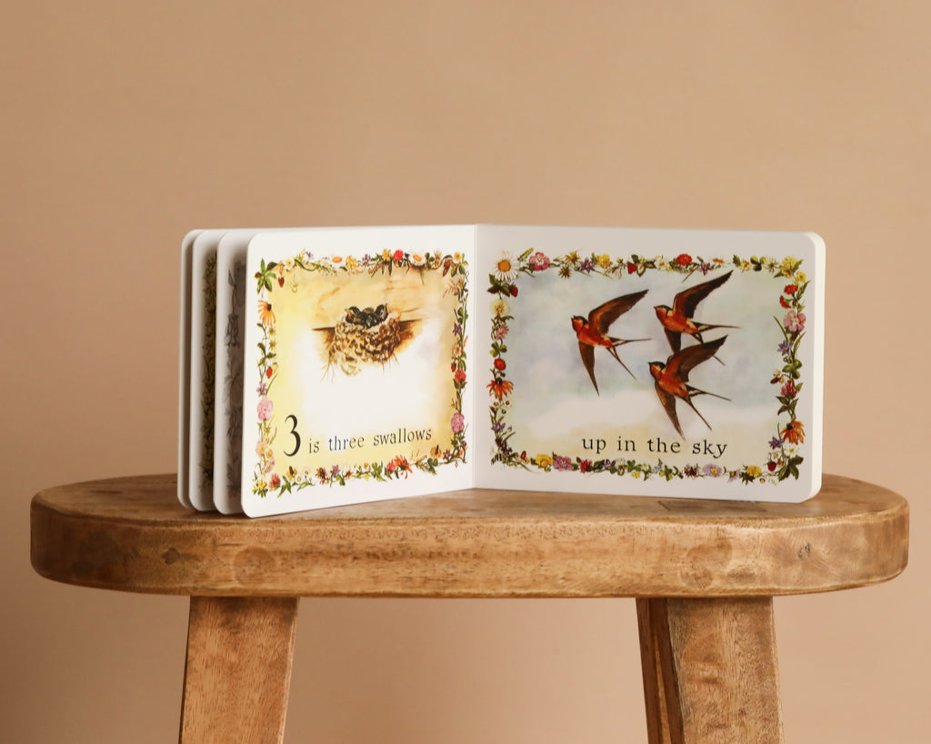 An open copy of "1 Is One by Tasha Tudor" rests on a wooden stool, displaying baby swallows in a nest and soaring in the sky—a charming counting board book with Tasha Tudor’s classic illustrative style.