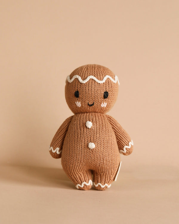 The Cuddle + Kind Baby Gingerbread is a collectible knitted doll made from Peruvian cotton yarn that stands upright. It showcases embroidered eyes and a smile, along with white icing details on its head, arms, and three buttons on its body against a plain beige background.