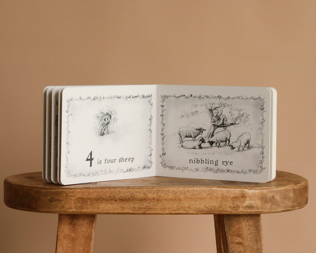 1 Is One by Tasha Tudor is open on a wooden stool, showing black-and-white illustrations and counting text. The classic board book rests against a plain beige background.