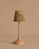 A small, peach-colored table lamp with a floral-patterned brown and white lampshade stands next to the Maileg Christmas Morning Living Room Set fireplace against a plain beige background.
