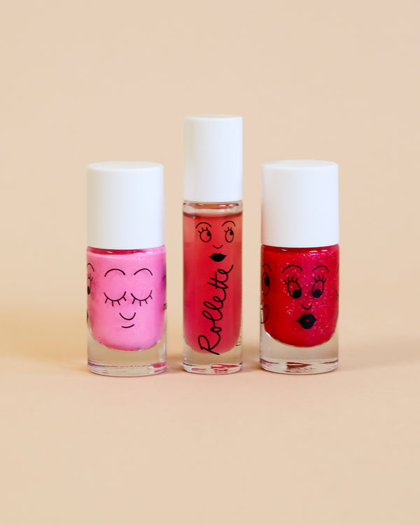 Three bottles of Nailmatic vegan cruelty-free nail polish with cartoon faces on them, standing in a row against a soft peach background. The bottles are in shades of pink and red, each decorated with glitter.