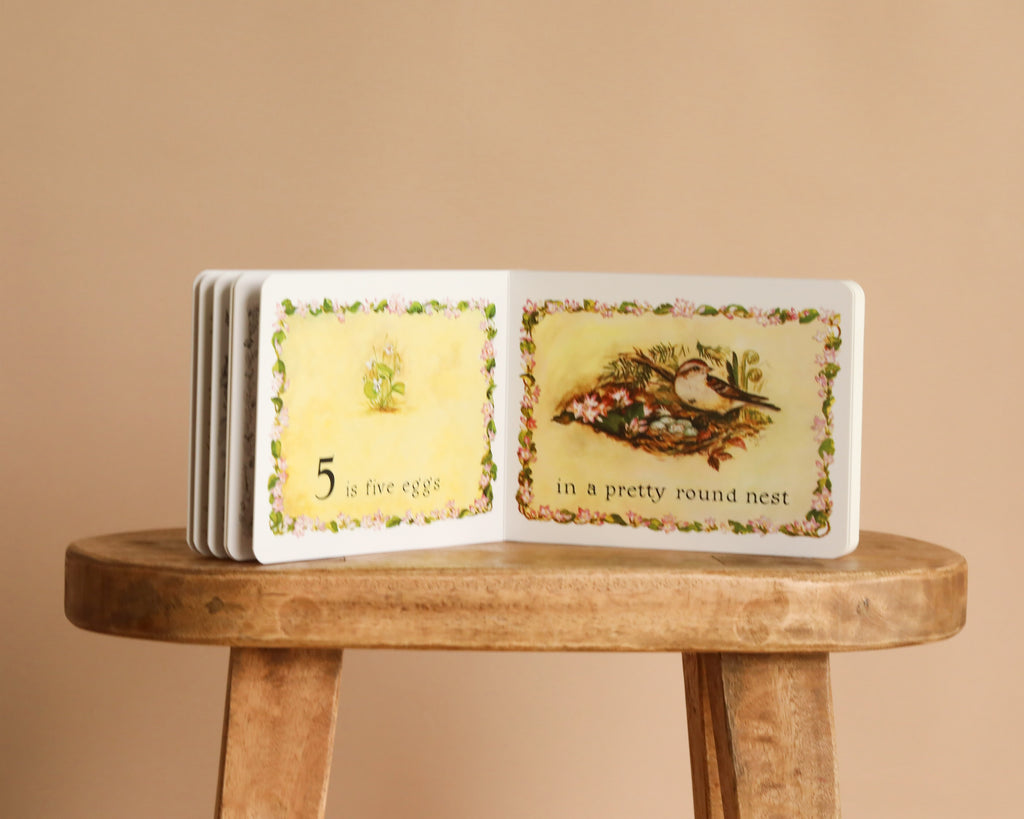 An open copy of *1 Is One by Tasha Tudor* rests on a wooden stool, displaying "5 is five eggs" and "in a pretty round nest"—a charming counting book for fans of Tasha Tudor’s classic style.