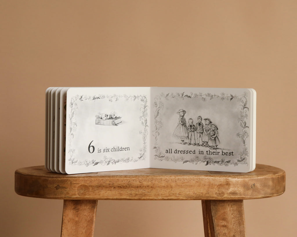 1 Is One by Tasha Tudor sits open on a wooden stool, displaying six children in fancy clothes. The left page reads, "6 is six children," and the right, "all dressed in their best"—a charming counting classic.