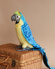 A vibrant blue and yellow Macaw Bird Stuffed Animal, realistically crafted as a stuffed animal, sits perched on a woven wicker basket against a neutral beige background. Its feathers are bright and its eyes alert.