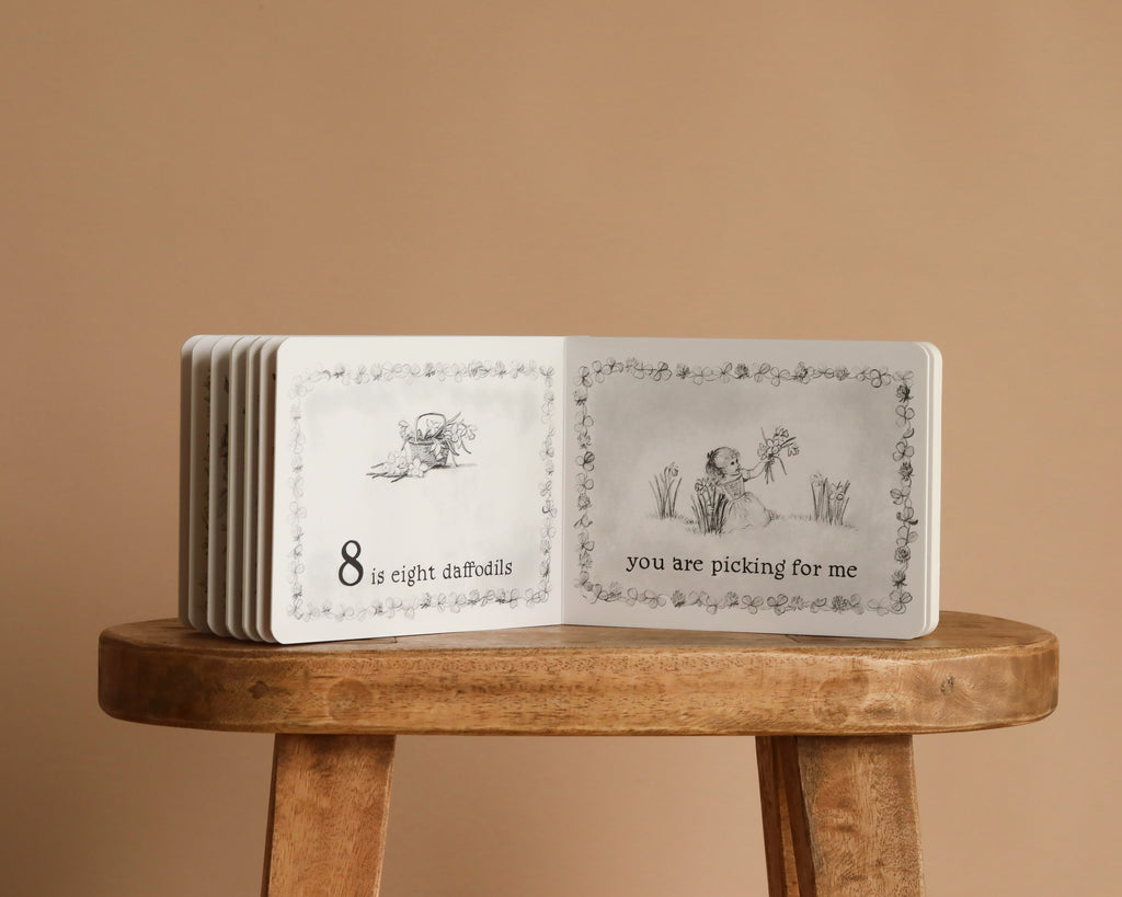 1 Is One by Tasha Tudor, a Classic Board Book, is open on a wooden stool, showing illustrations of daffodils and a girl picking flowers. The charming counting book reads, "8 is eight daffodils," and "you are picking for me.