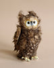 A Owl Stuffed Animal with fluffy brown feathers, large expressive blue eyes, and a quizzical expression. The owl is standing upright on its yellow feet against a plain beige background. This high-quality plush
