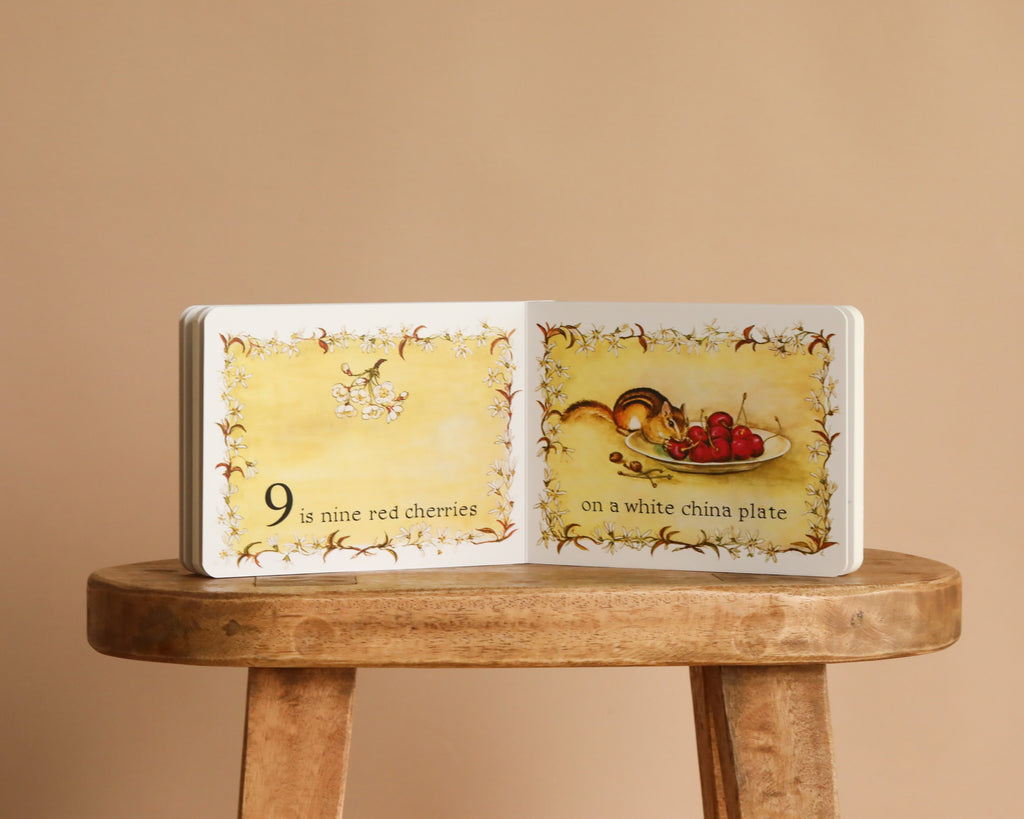 1 Is One by Tasha Tudor is open on a wooden stool. The left page shows the number 9 and cherries, with “9 is nine red cherries.” The right page shows a plate of cherries and the text "on a white china plate." A charming counting book!.