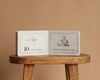 An open copy of "1 Is One" by Tasha Tudor rests on a wooden stool against a beige background, displaying a charming counting scene with a boy writing, and text including “10 is ten numbers” and “Tom has written on his slate.”.