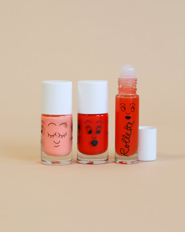 Three bottles of Nailmatic - 2 Nail Polish + 1 Lipgloss Set - Amazing Trip with cute, cartoonish faces printed on them, displayed against a warm beige background. The center bottle has red polish and the other two are pink.