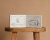 An open copy of “1 Is One” by Tasha Tudor rests on a wooden stool, displaying an illustration of twelve baby birds with the text: "12 is twelve baby birds learning how to sing," set against a charming beige background.