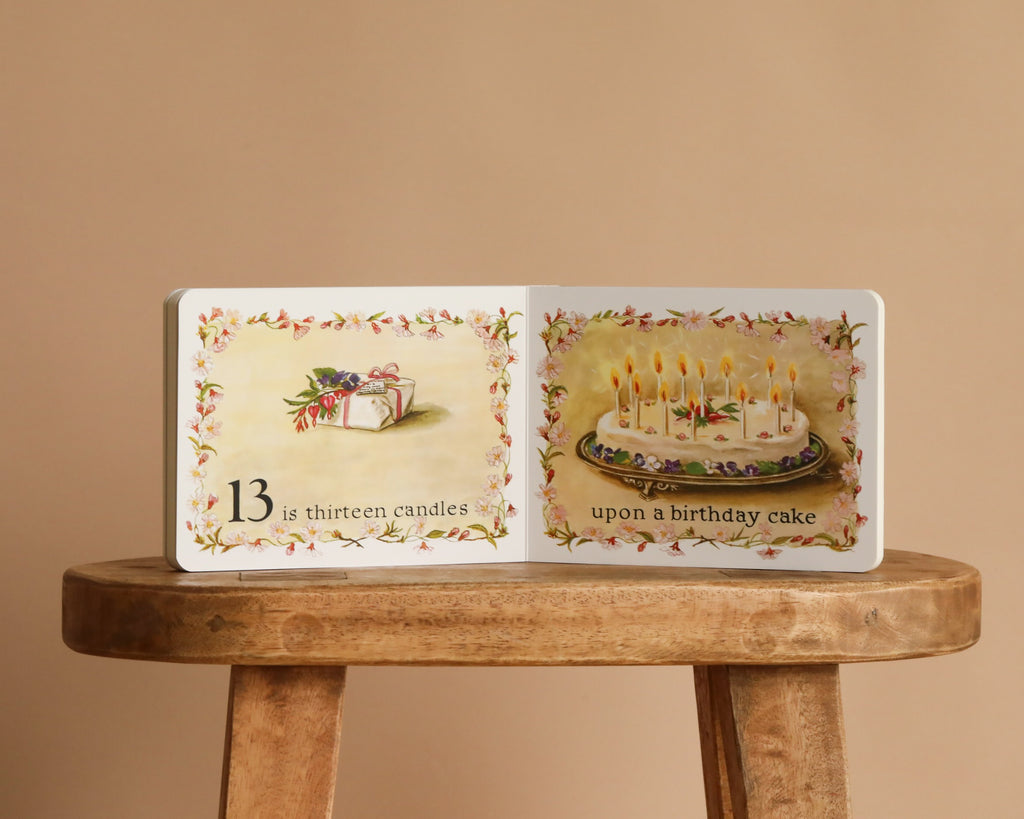 An open copy of "1 Is One" by Tasha Tudor sits on a wooden stool, showing an illustrated gift box with flowers and a cake with thirteen candles. The page says: "13 is thirteen candles upon a birthday cake." Classic Board Book style.