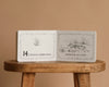 An open copy of "1 Is One" by Tasha Tudor rests on a wooden stool, turned to the page displaying "14 is fourteen mallard ducks swimming on a lake," with an illustration of ducks in water from this classic counting book.