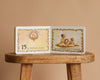 1 Is One by Tasha Tudor, a classic board book, rests open on a wooden stool, showing “15 is fifteen roses” with a wreath and two girls making a rose wreath—charmingly illustrated with floral borders throughout.
