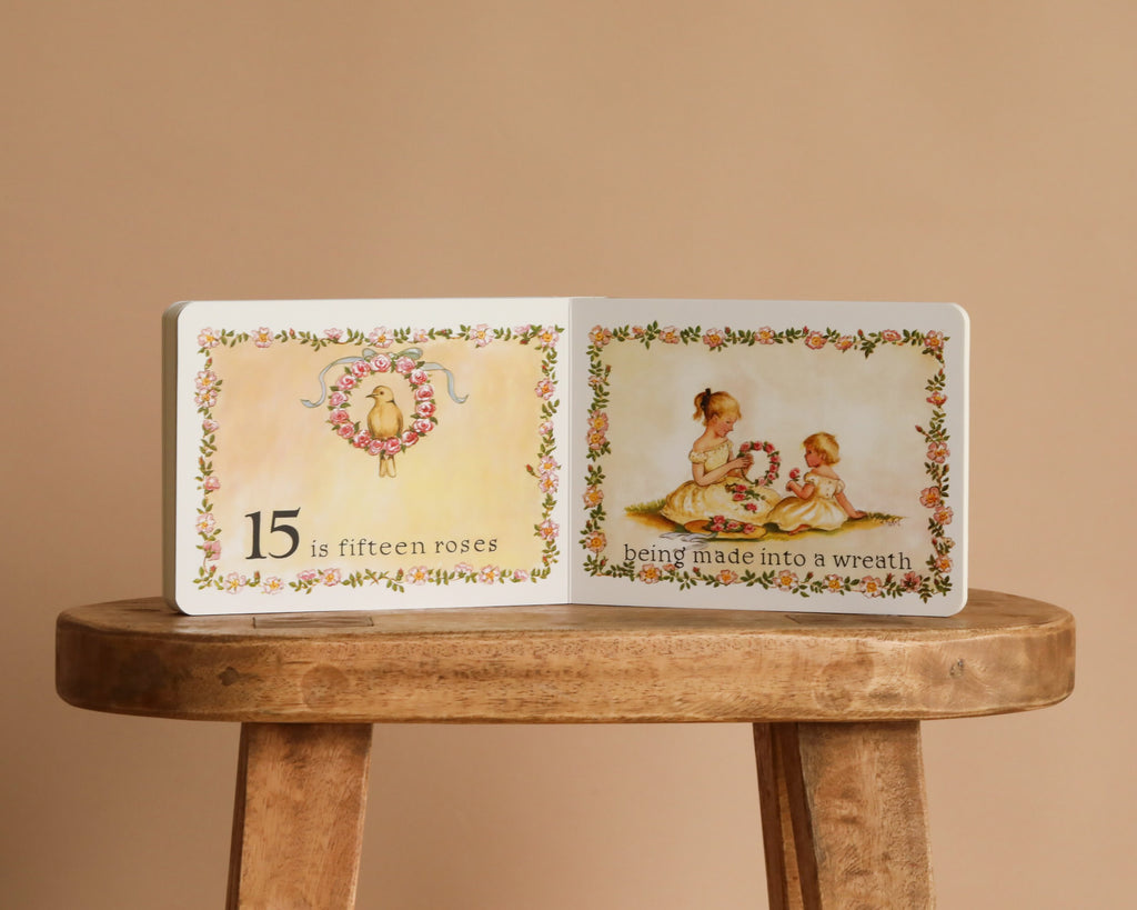 1 Is One by Tasha Tudor, a classic board book, rests open on a wooden stool, showing “15 is fifteen roses” with a wreath and two girls making a rose wreath—charmingly illustrated with floral borders throughout.