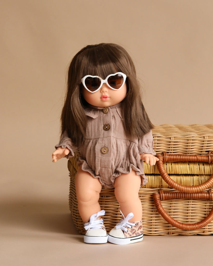 The Minikane Baby Doll (13") - Cherry with Clothing features long brown hair, white heart-shaped sunglasses, a brown romper, and sneakers, posed in front of a woven basket on a beige background. This anatomically correct doll is made in Spain.