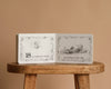 A classic counting book, "1 Is One by Tasha Tudor," sits open on a wooden stool. Pencil illustrations show “18 is eighteen stars” and “twinkling in the sky,” all on a beige background in Tudor's signature style.