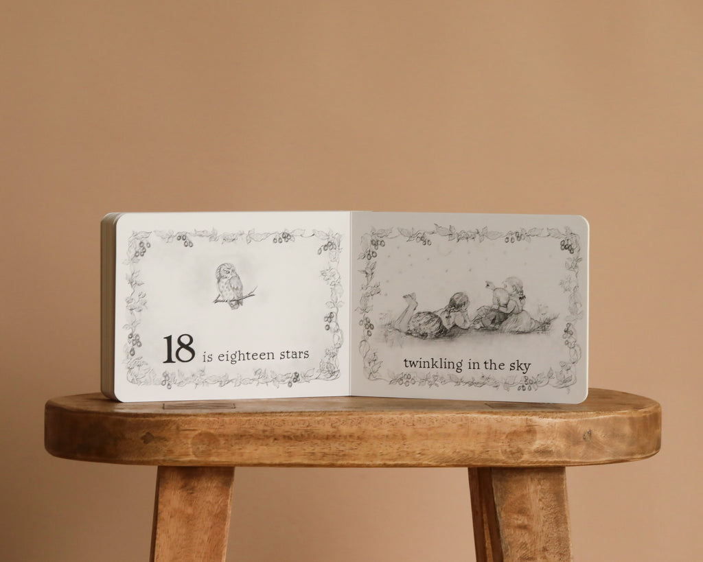 A classic counting book, "1 Is One by Tasha Tudor," sits open on a wooden stool. Pencil illustrations show “18 is eighteen stars” and “twinkling in the sky,” all on a beige background in Tudor's signature style.