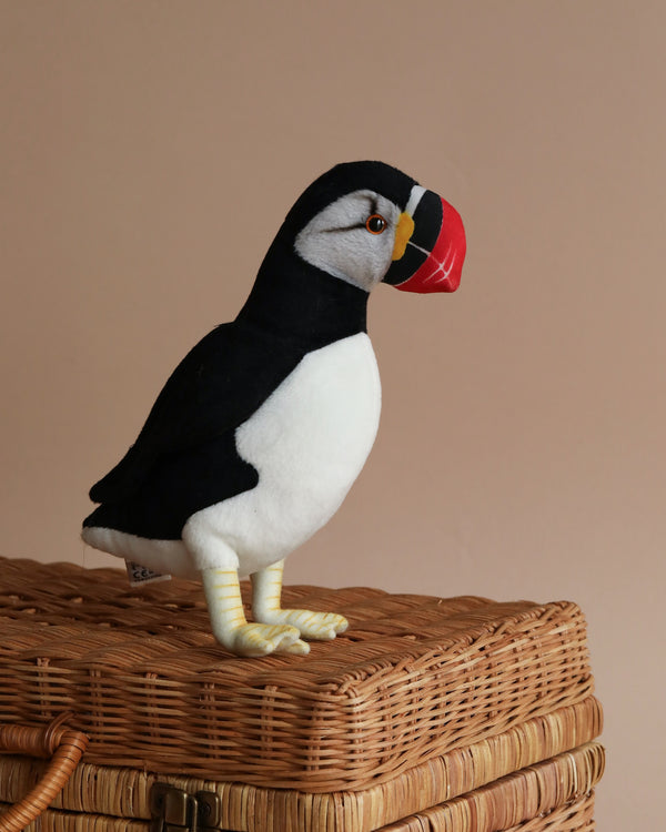 A Puffin Bird Stuffed Animal with black and white feathers and a colorful beak, standing on a wicker basket against a pale pink background.