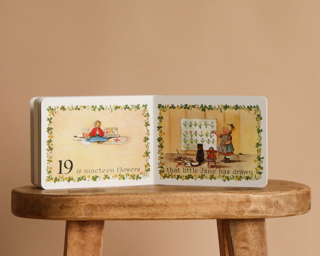 An open copy of 1 Is One by Tasha Tudor sits on a wooden stool, showing a girl drawing flowers and sharing them with her cat, while the text counts nineteen flowers in Tasha Tudor’s signature enchanting style.