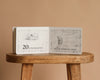An open copy of "1 Is One" by Tasha Tudor rests on a wooden stool. One page shows a goose with the text "20 is twenty geese," while the opposite page has a girl watching a flock flying at dawn.