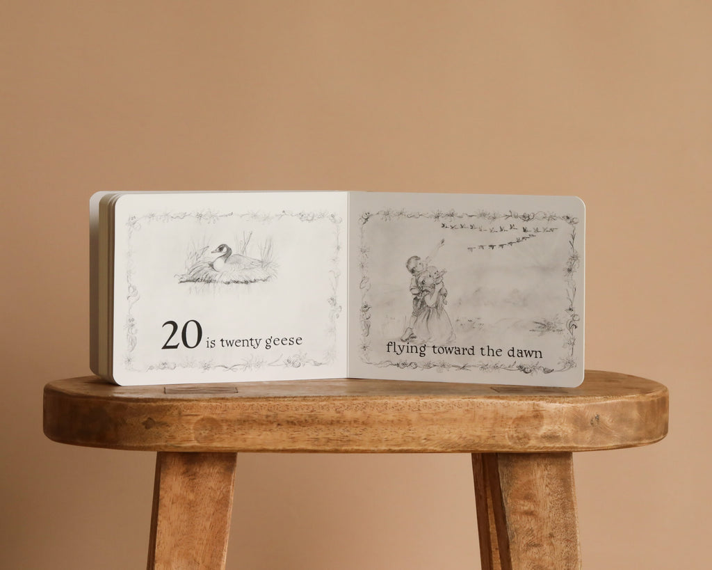 An open copy of "1 Is One" by Tasha Tudor rests on a wooden stool. One page shows a goose with the text "20 is twenty geese," while the opposite page has a girl watching a flock flying at dawn.