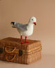 A realistic Seagull Bird Stuffed Animal with a white and gray body and red beak standing on top of a woven wicker basket against a plain light brown background.