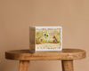 The board book “1 Is One by Tasha Tudor” stands upright on a wooden stool against a plain beige background, with its cover showing a child and rabbit surrounded by greenery and flowers.