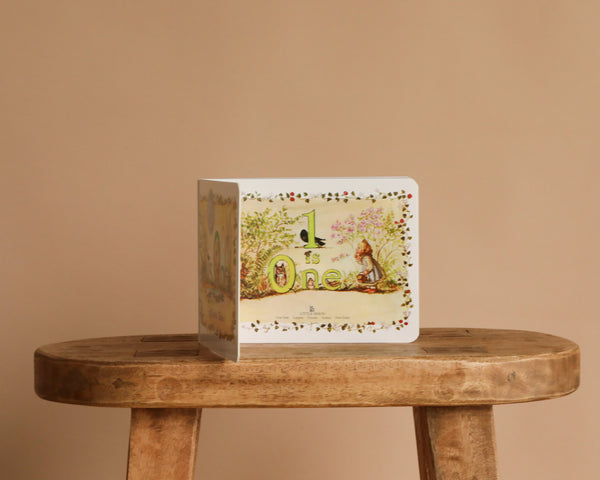 The board book “1 Is One by Tasha Tudor” stands upright on a wooden stool against a plain beige background, with its cover showing a child and rabbit surrounded by greenery and flowers.