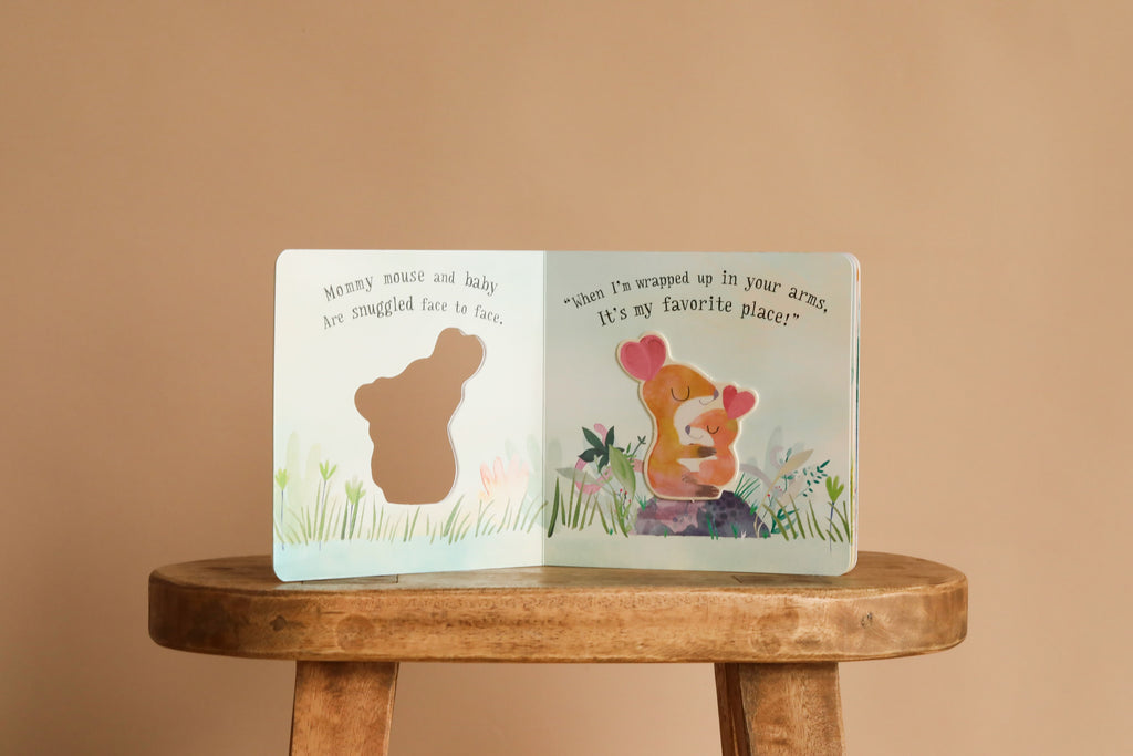 An open copy of "I Love You So" by Patricia Hegarty rests on a wooden stool. The left page features a mouse cut-out with, "Mommy mouse and baby are snuggled face to face," while the right shows baby animals hugging: "It’s my favorite place!.